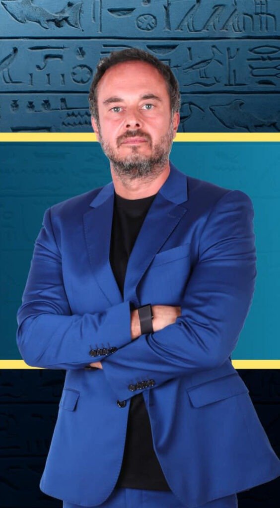 Male portrait in a blue suit with arms crossed, standing before a teal backdrop with hieroglyphic patterns.